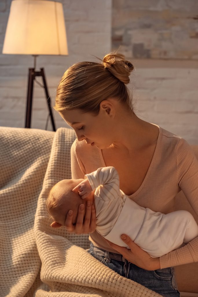 young mother sitting on sofa and holding newborn baby during breastfeeding at night